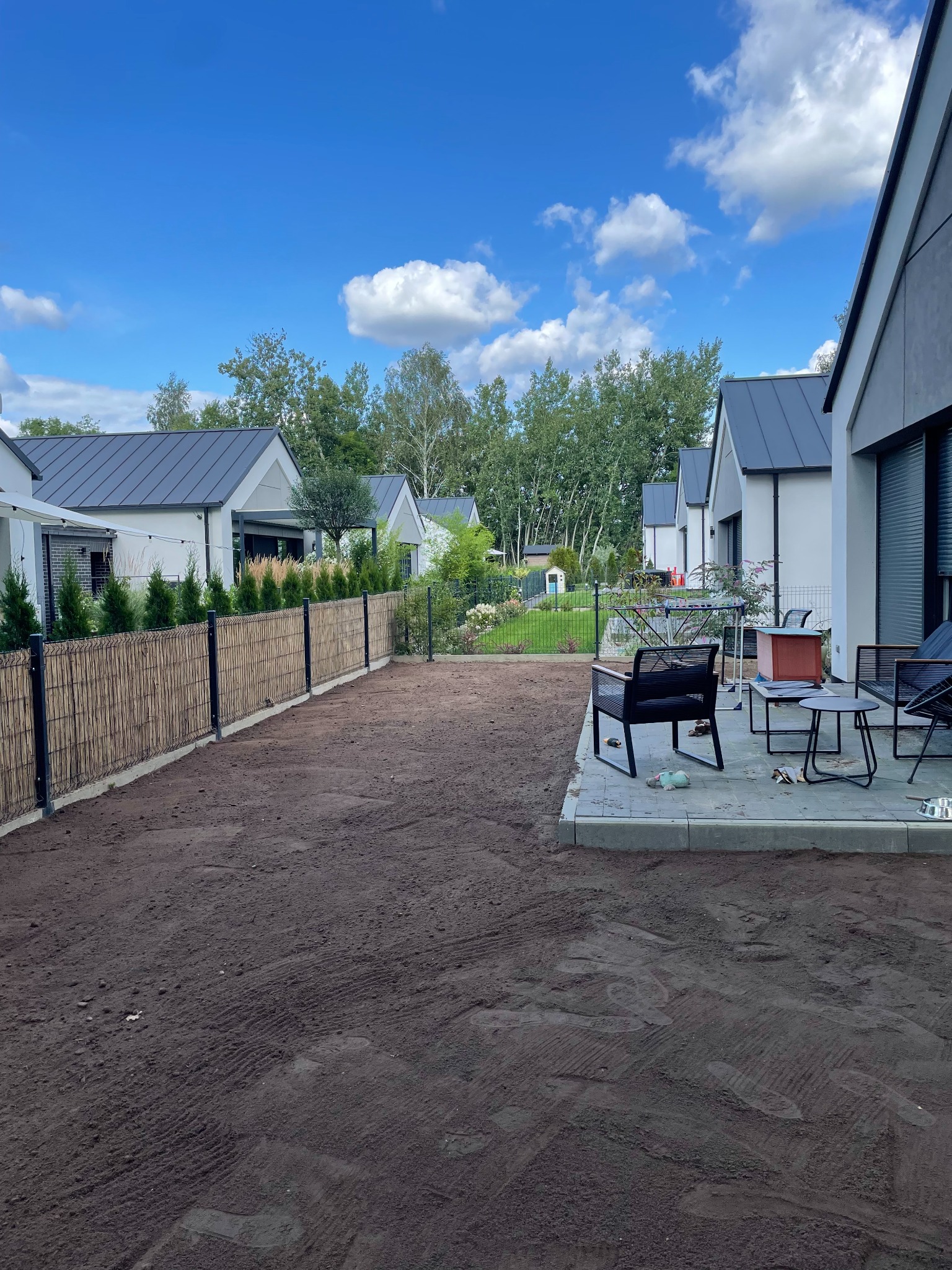 Przygotowany teren pod trawnik, wyrównana ziemia z widocznymi śladami maszyn, nowoczesne domy w tle, ogrodzenie z bambusa i metalu, taras z meblami ogrodowymi.