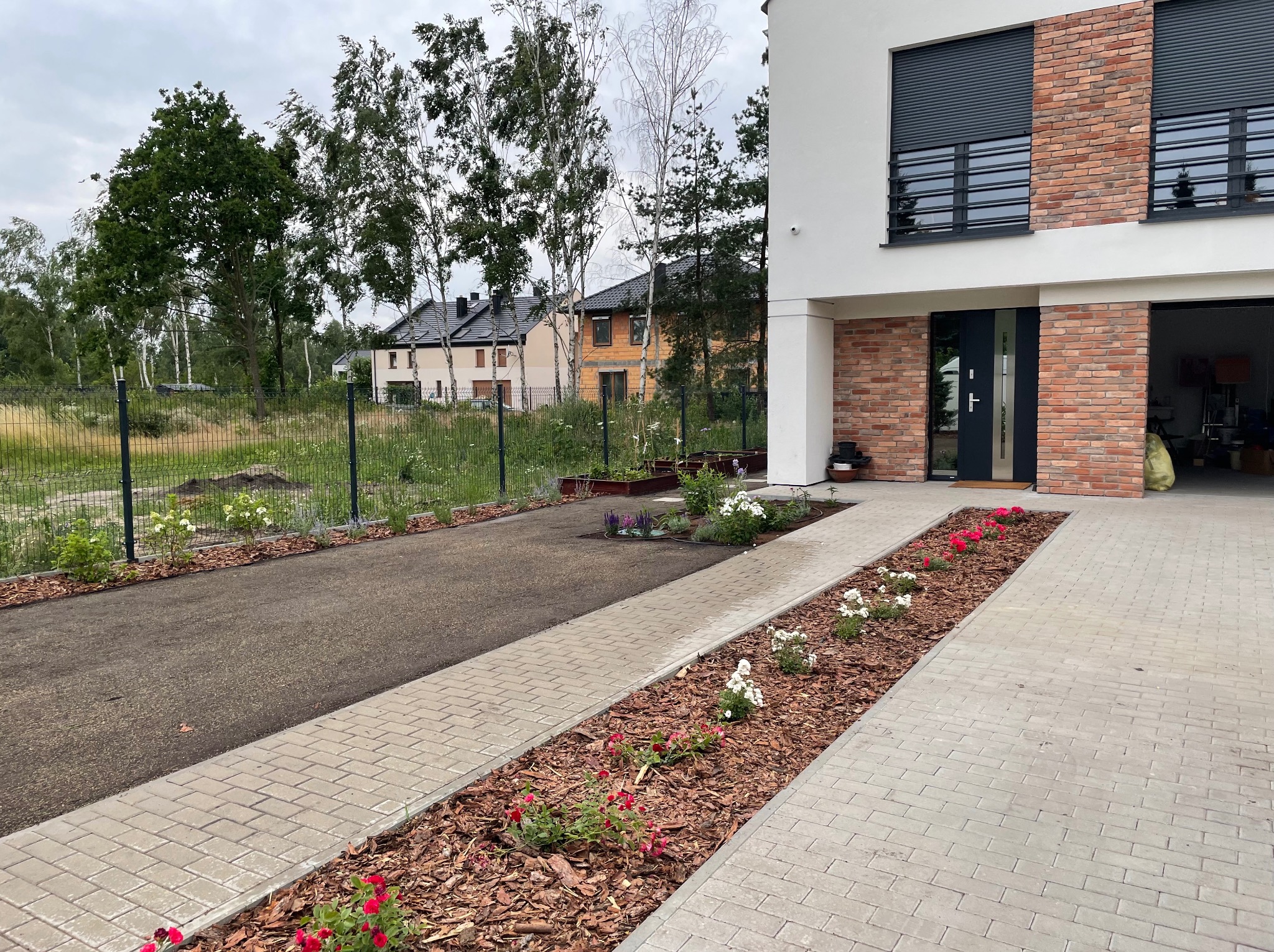 Nowoczesny dom z podjazdem wyłożonym kostką brukową i pasem ziemi z nasadzeniami kwiatów oraz kory, widok z poziomu gruntu.