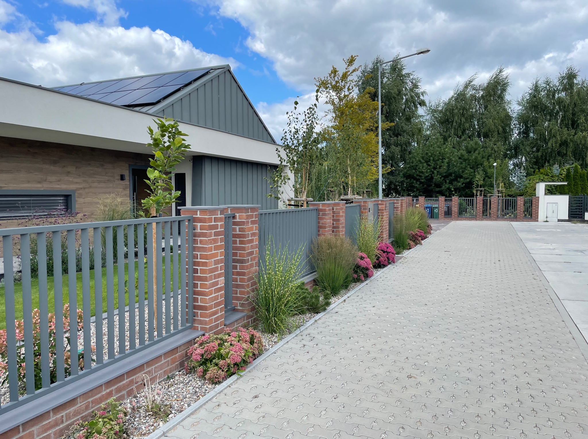 Nowoczesny dom z panelami słonecznymi, szarym ogrodzeniem z ceglanymi słupkami i brukowaną ścieżką z ozdobnymi roślinami wzdłuż ogrodzenia.