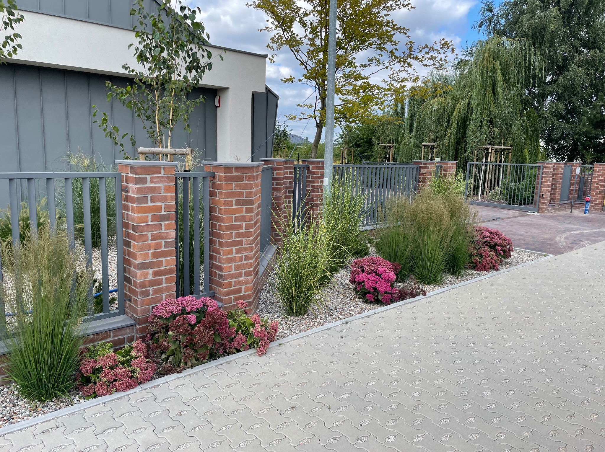 Nowoczesny ogród przydomowy z szarą metalową bramą, ceglanymi słupkami, ozdobnymi trawami, różowymi kwiatami i jasnoszarą kostką brukową.