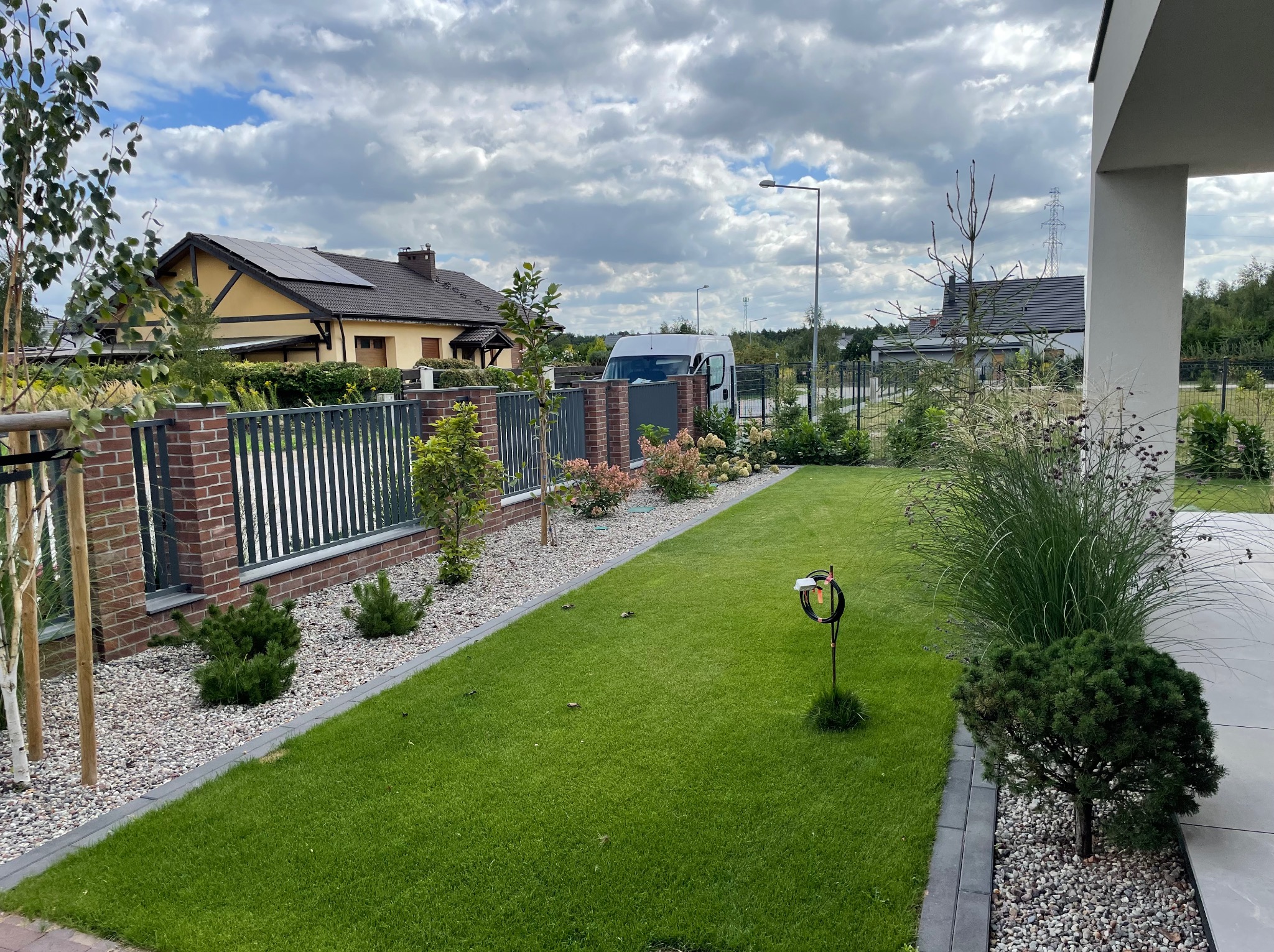 Zadbany trawnik z systemem nawadniania, obrzeżami z kostki, rabatami wysypanymi kamieniem, młodymi drzewkami i krzewami, ogrodzeniem z cegły i metalu, na tle domów i pochmurnego nieba.