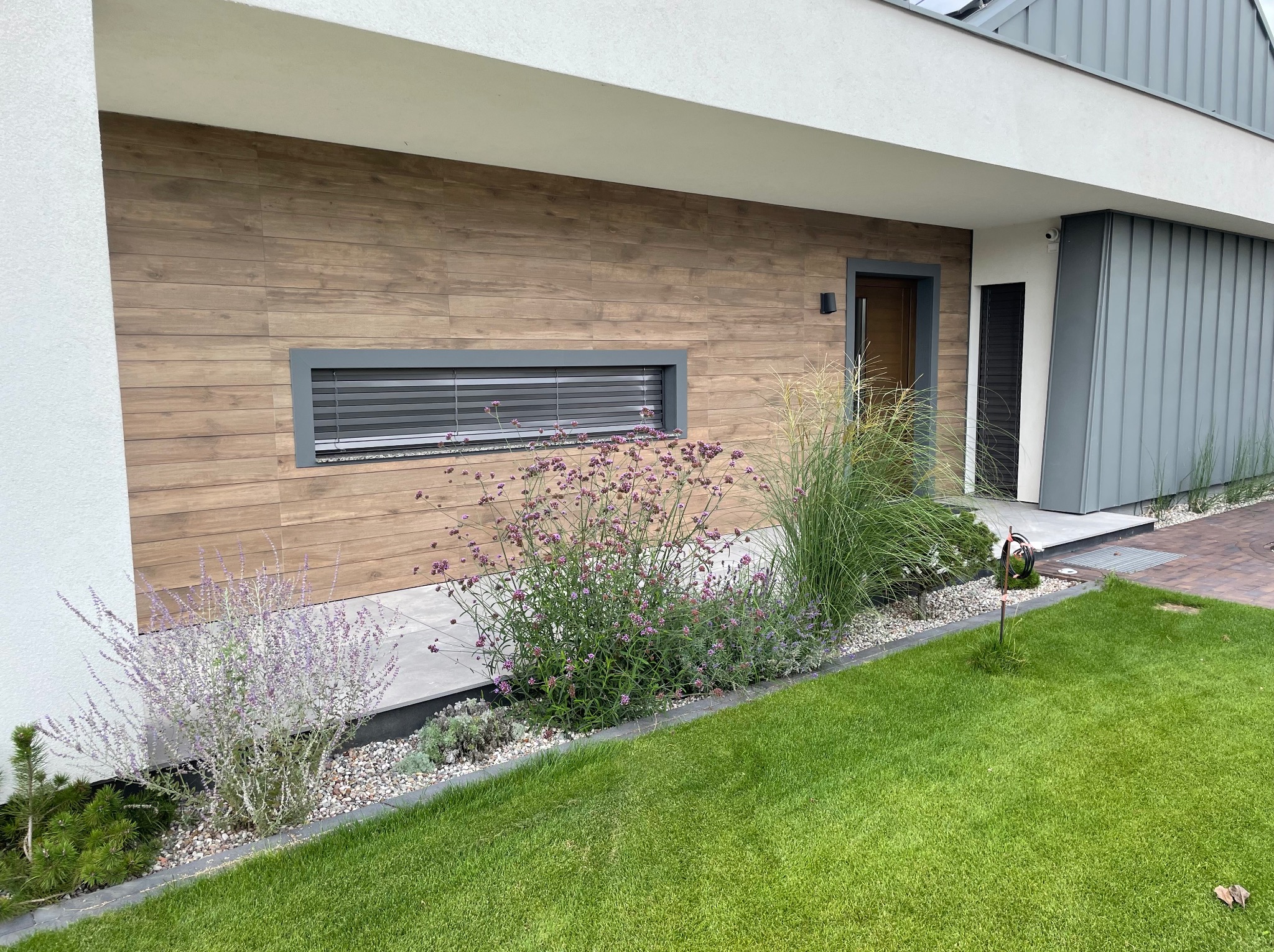 Nowoczesny ogród przydomowy z rabatami kwiatowymi, ozdobnymi trawami i równo przystrzyżonym trawnikiem przed budynkiem z elewacją imitującą drewno.