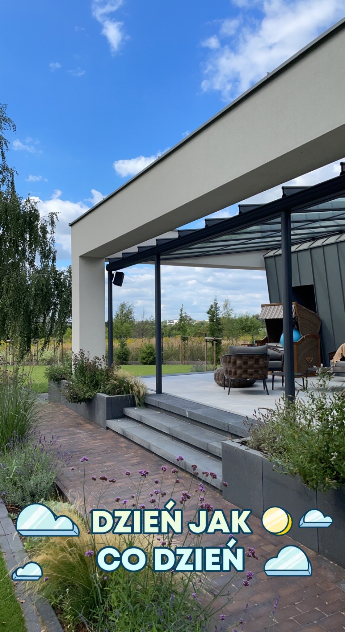 Nowoczesny taras z zadaszeniem, meblami ogrodowymi i zielenią, widok na zadbany trawnik oraz błękitne niebo z chmurami.