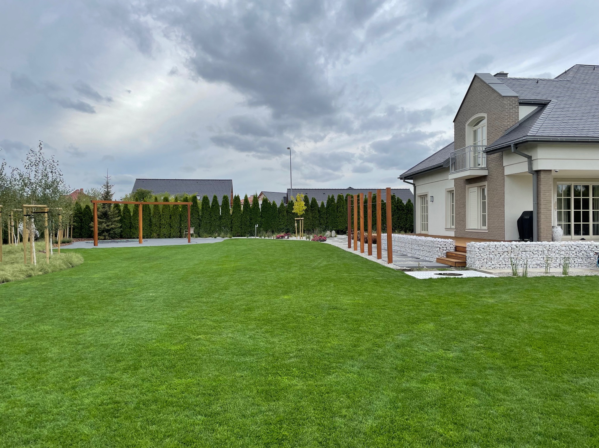 Rozległy, zielony trawnik przed nowoczesnym domem z elementami gabionowymi, drewnianymi pergolami i równo przyciętym żywopłotem z tui.