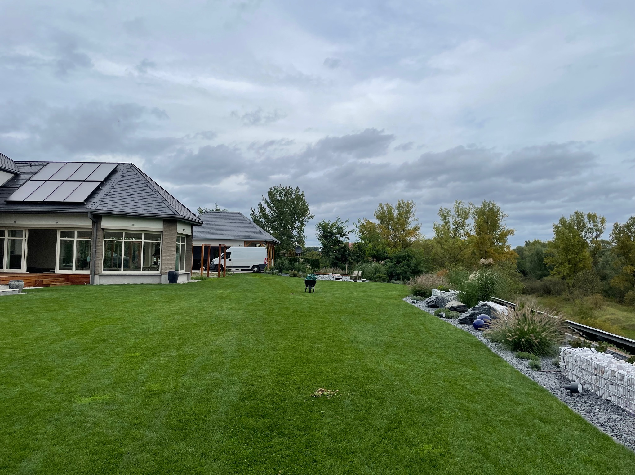 Rozległy, zielony trawnik przed nowoczesnym domem z panelami słonecznymi na dachu, z widoczną taczką ogrodową i elementami dekoracyjnymi w postaci kamieni i roślin ozdobnych.