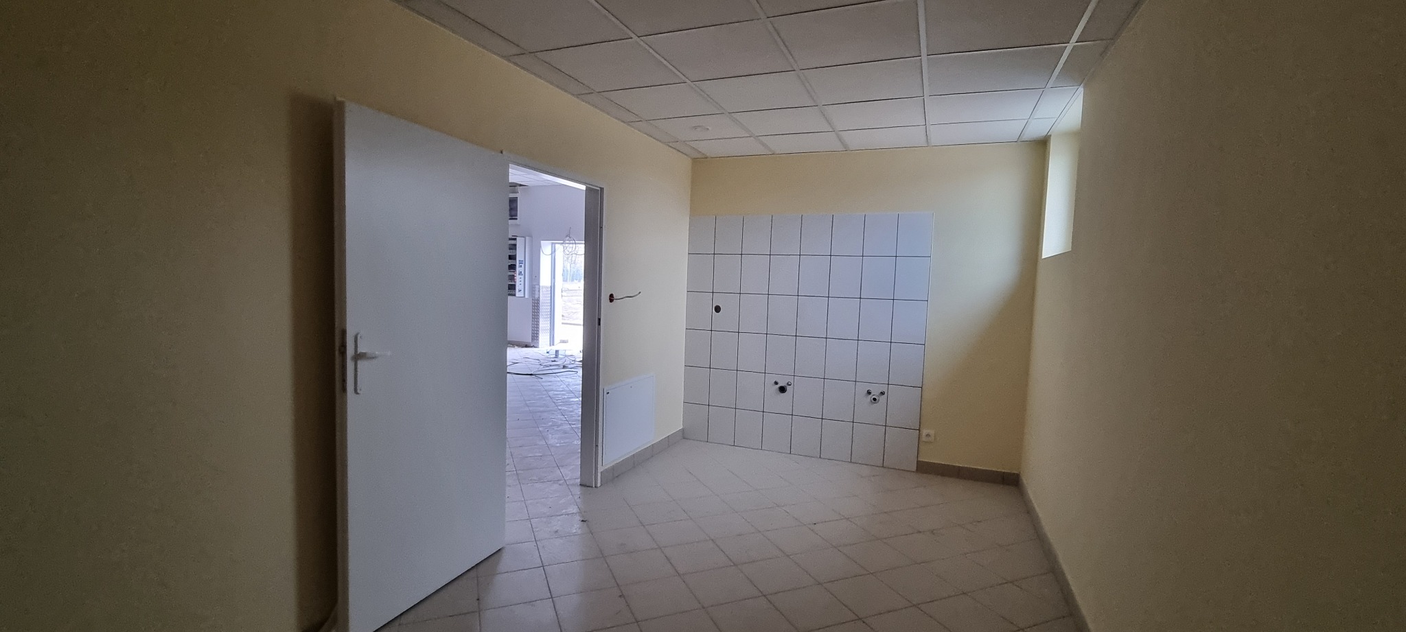 Pusta, jasna przestrzeń z otwartymi drzwiami, kwadratowymi płytkami na ścianie i suficie kasetonowym, przygotowana do dalszych prac wykończeniowych.