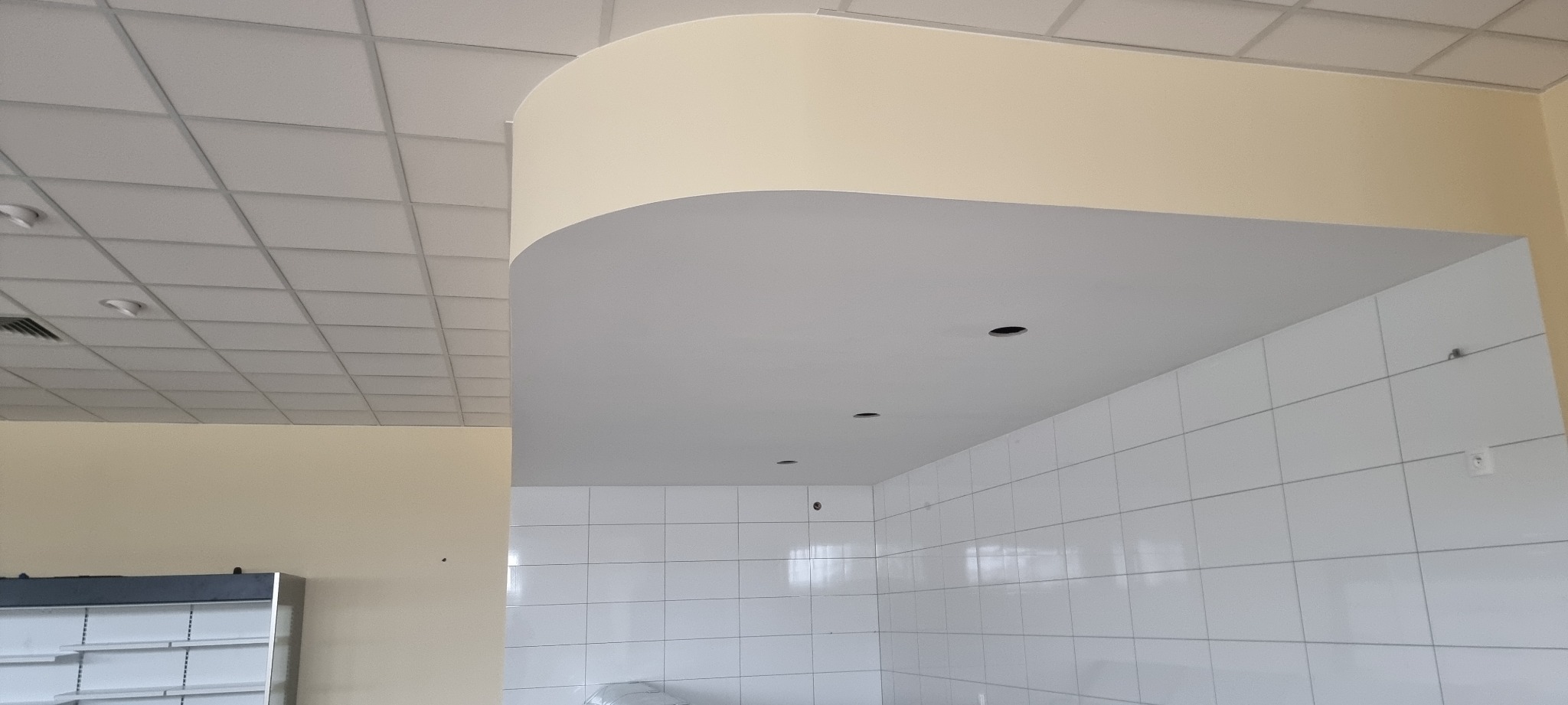 Wnętrze lokalu w trakcie wykańczania: widoczny sufit podwieszany, fragment ściany pomalowanej na kremowo oraz białe płytki na ścianie z przygotowanymi otworami na oświetlenie.