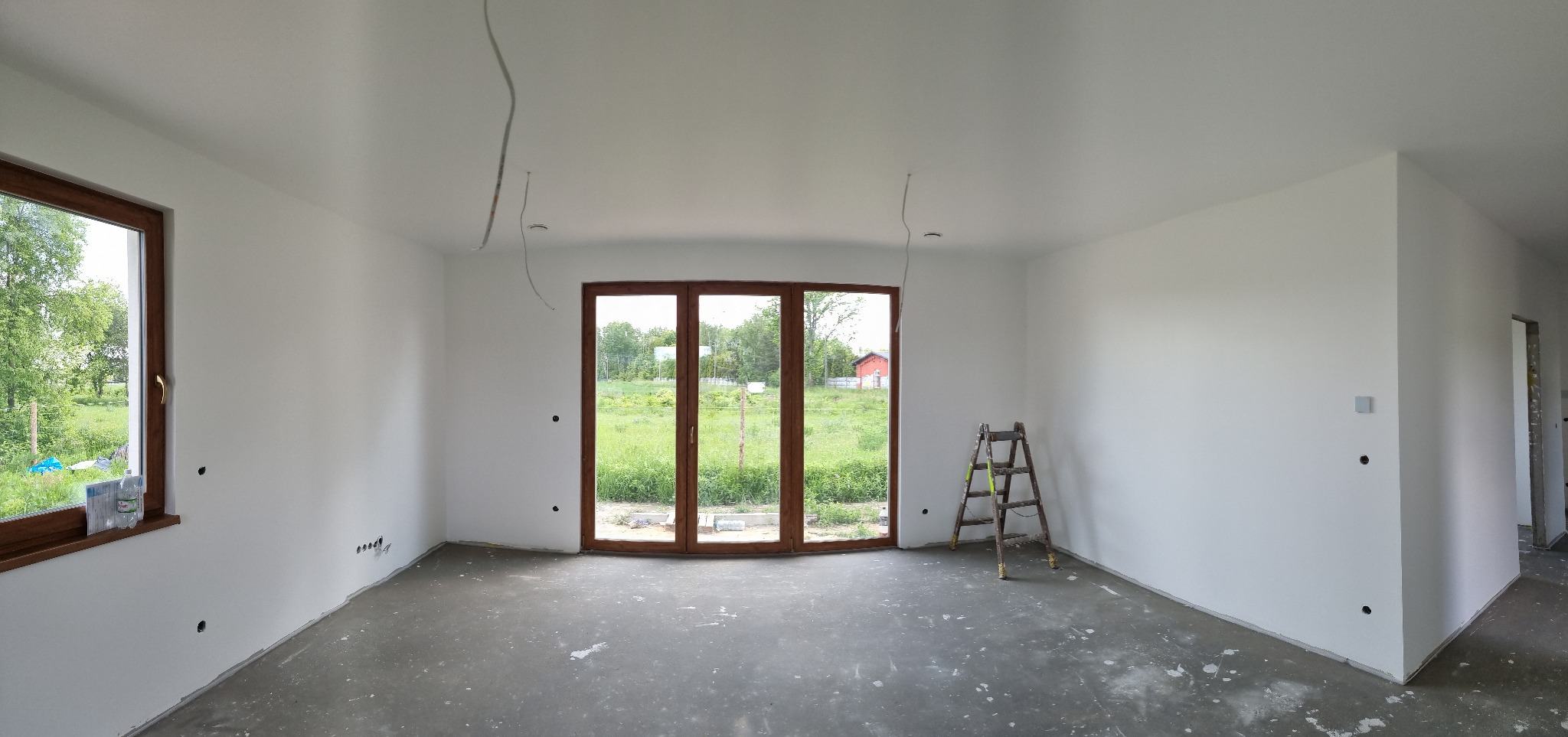 Jasne, świeżo pomalowane wnętrze z drewnianą stolarką okienną i drabiną. Widoczne przygotowane punkty elektryczne i surowa podłoga. Duże okno z widokiem na zieleń.