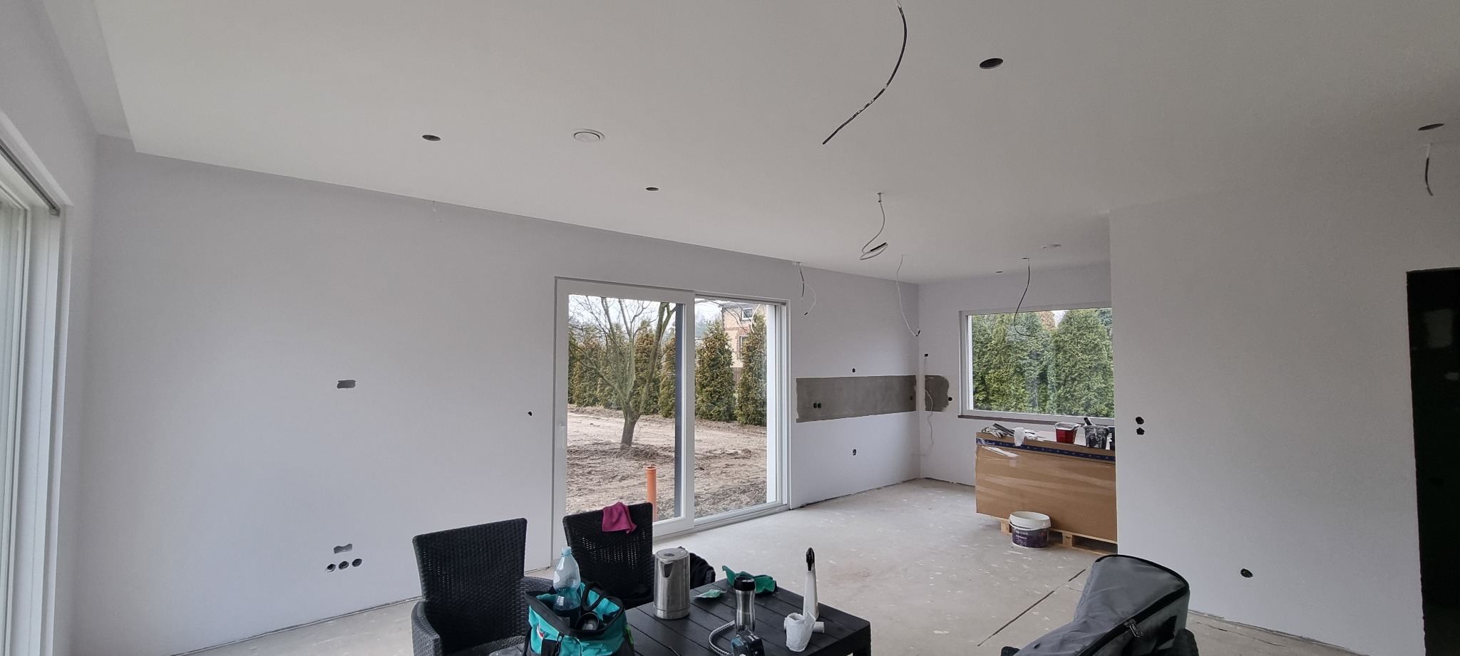 Jasne wnętrze po szpachlowaniu i malowaniu ścian na biało, z widocznymi przewodami elektrycznymi i oknami z widokiem na zieleń. Przygotowane do dalszych prac wykończeniowych.