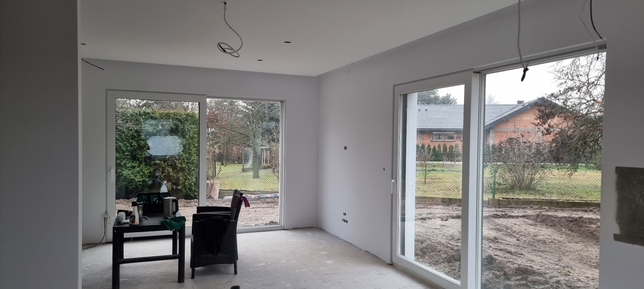 Jasne, świeżo pomalowane wnętrze z dużymi oknami przesuwnymi, przez które widać ogród. Widoczne ślady po gniazdkach elektrycznych i wiszące kable oświetleniowe.