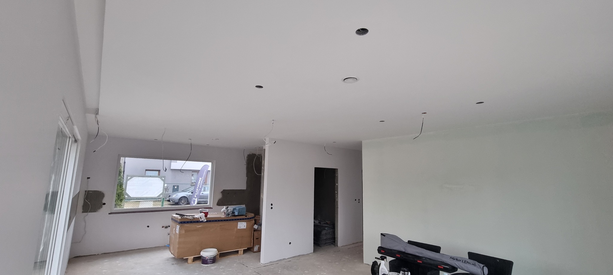 Wnętrze w trakcie prac wykończeniowych: świeżo pomalowane ściany na biało, widoczne otwory na oświetlenie w suficie, narzędzia i materiały budowlane przy oknie.