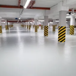 Jasnoszara posadzka żywiczna w garażu podziemnym, z rzędami słup&oacute;w oznaczonych czarno-ż&oacute;łtymi pasami, widoczne linie wyznaczające miejsca parkingowe.