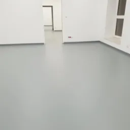 Gładka, szara podłoga w jasnym pomieszczeniu z białymi ścianami i widocznymi gniazdkami elektrycznymi przy podłodze, z otwartym przejściem do kolejnego pomieszczenia w tle.