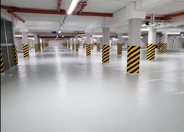 Jasnoszara posadzka żywiczna w garażu podziemnym, z rzędami słupów oznaczonych czarno-żółtymi pasami, widoczne linie wyznaczające miejsca parkingowe.