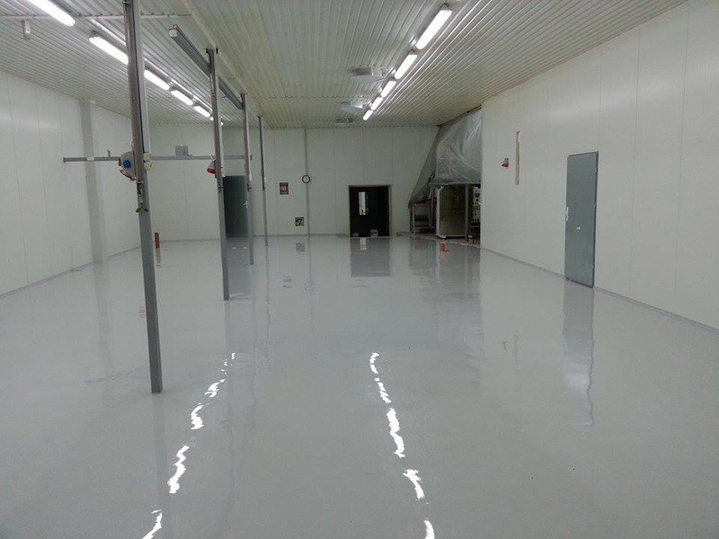 Jasnoszara, błyszcząca posadzka przemysłowa w hali produkcyjnej z metalowymi słupami i białymi ścianami panelowymi.