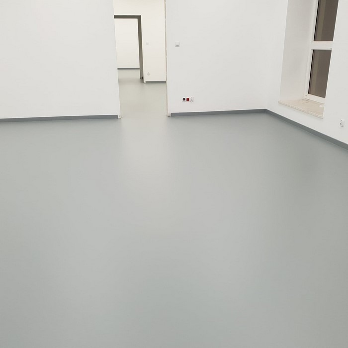 Gładka, szara podłoga w jasnym pomieszczeniu z białymi ścianami i widocznymi gniazdkami elektrycznymi przy podłodze, z otwartym przejściem do kolejnego pomieszczenia w tle.