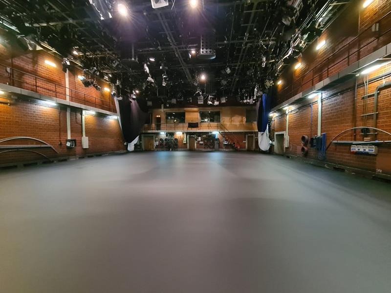 Przestronne studio telewizyjne z szarą, gładką posadzką, oświetleniem technicznym pod sufitem i ceglanymi ścianami.