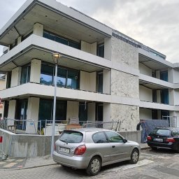 TSM - Budynek wielopiętrowy w trakcie wykończenia elewacji, z widocznymi fragmentami surowego betonu i zamontowanymi panelami elewacyjnymi o wzorze marmuru.