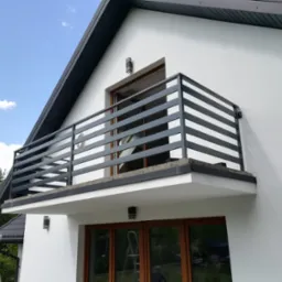 Nowoczesna, pozioma balustrada balkonowa w kolorze grafitowym na tle białej elewacji domu z drewnianą stolarką okienną i błękitnym niebem.