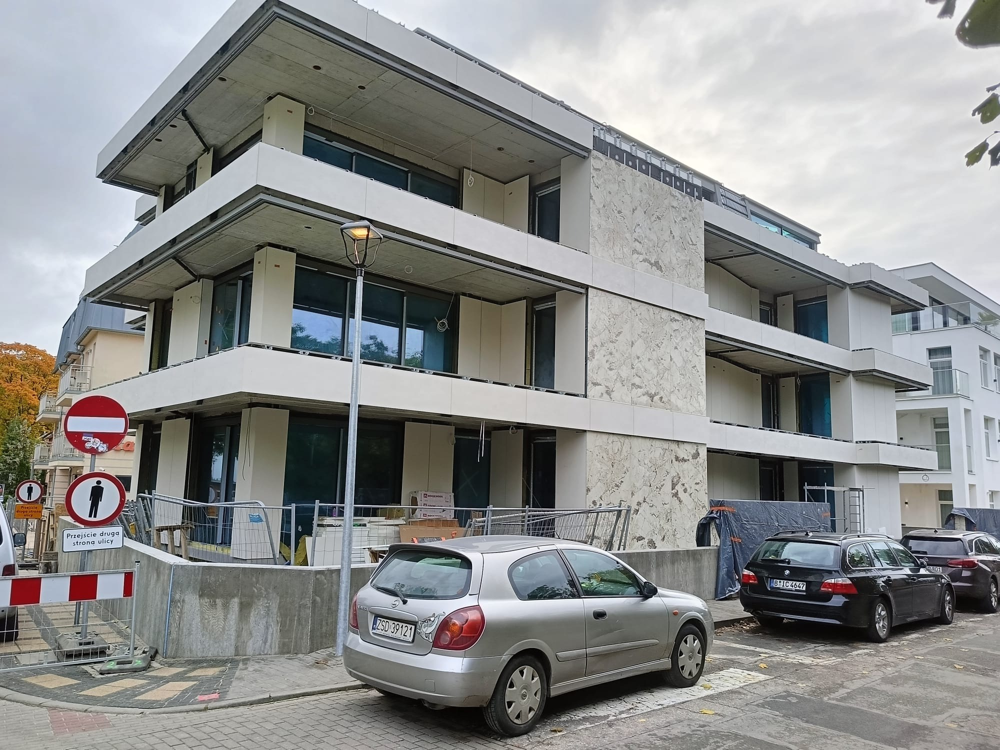 Budynek wielopiętrowy w trakcie wykończenia elewacji, z widocznymi fragmentami surowego betonu i zamontowanymi panelami elewacyjnymi o wzorze marmuru.