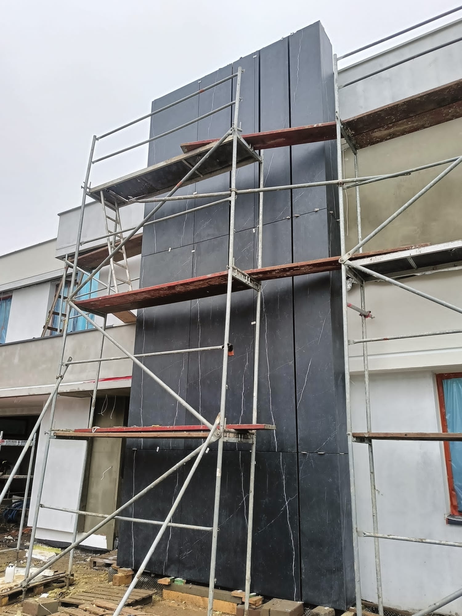 Montaż elewacji z płyt imitujących marmur, widoczne rusztowania. Elewacja w trakcie prac, płyty z wyraźnym wzorem białych żył na ciemnym tle.