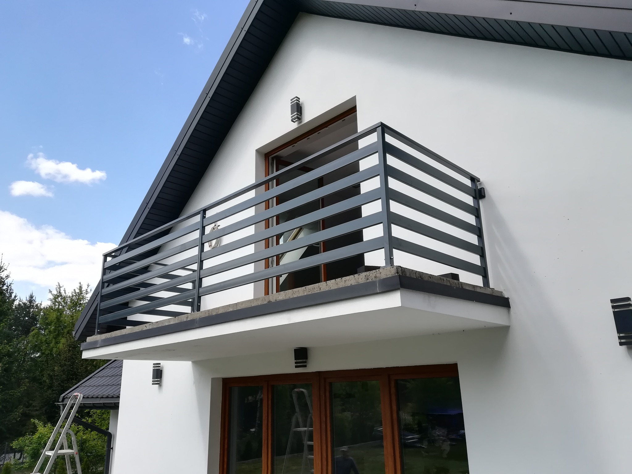 Nowoczesna, pozioma balustrada balkonowa w kolorze grafitowym na tle białej elewacji domu z drewnianą stolarką okienną i błękitnym niebem.