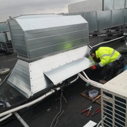 VENTIMAR SPÓŁKA Z OGRANICZONĄ ODPOWIEDZIALNOŚCIĄ - Pracownik w żółtej kurtce montuje metalowy kanał wentylacyjny na dachu budynku, widoczne narzędzia i kable.