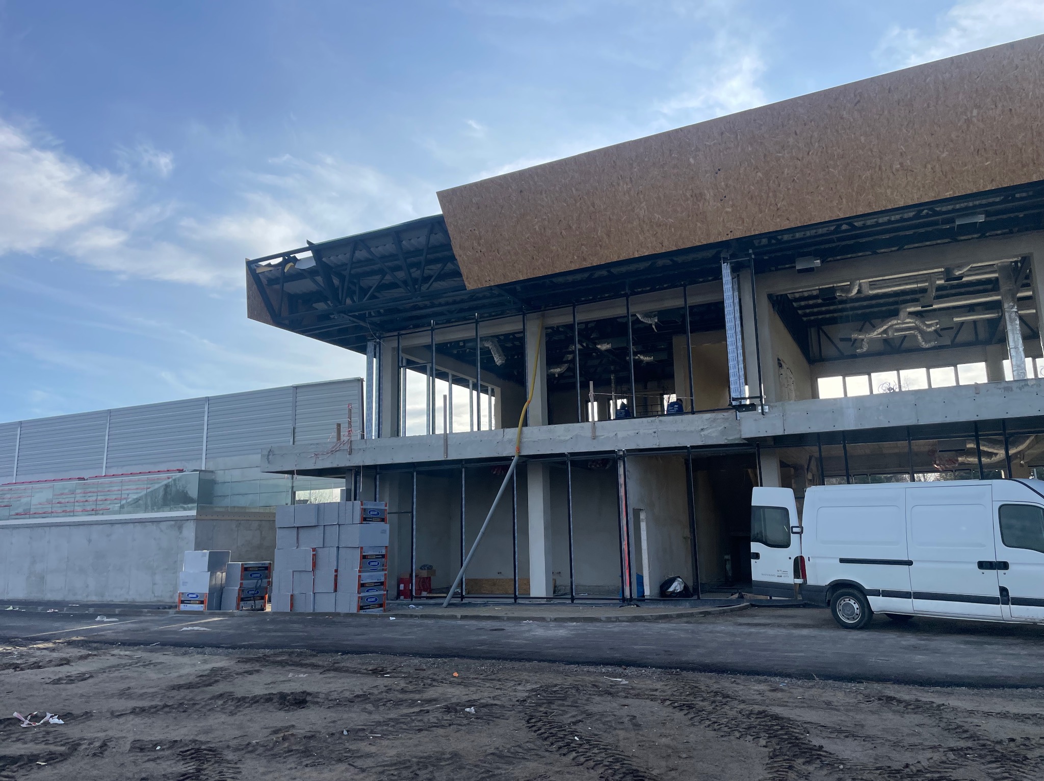 Budynek komercyjny w stanie surowym otwartym, widoczne elementy konstrukcyjne, instalacje wentylacyjne pod sufitem, palety z materiałami budowlanymi i biały van dostawczy.