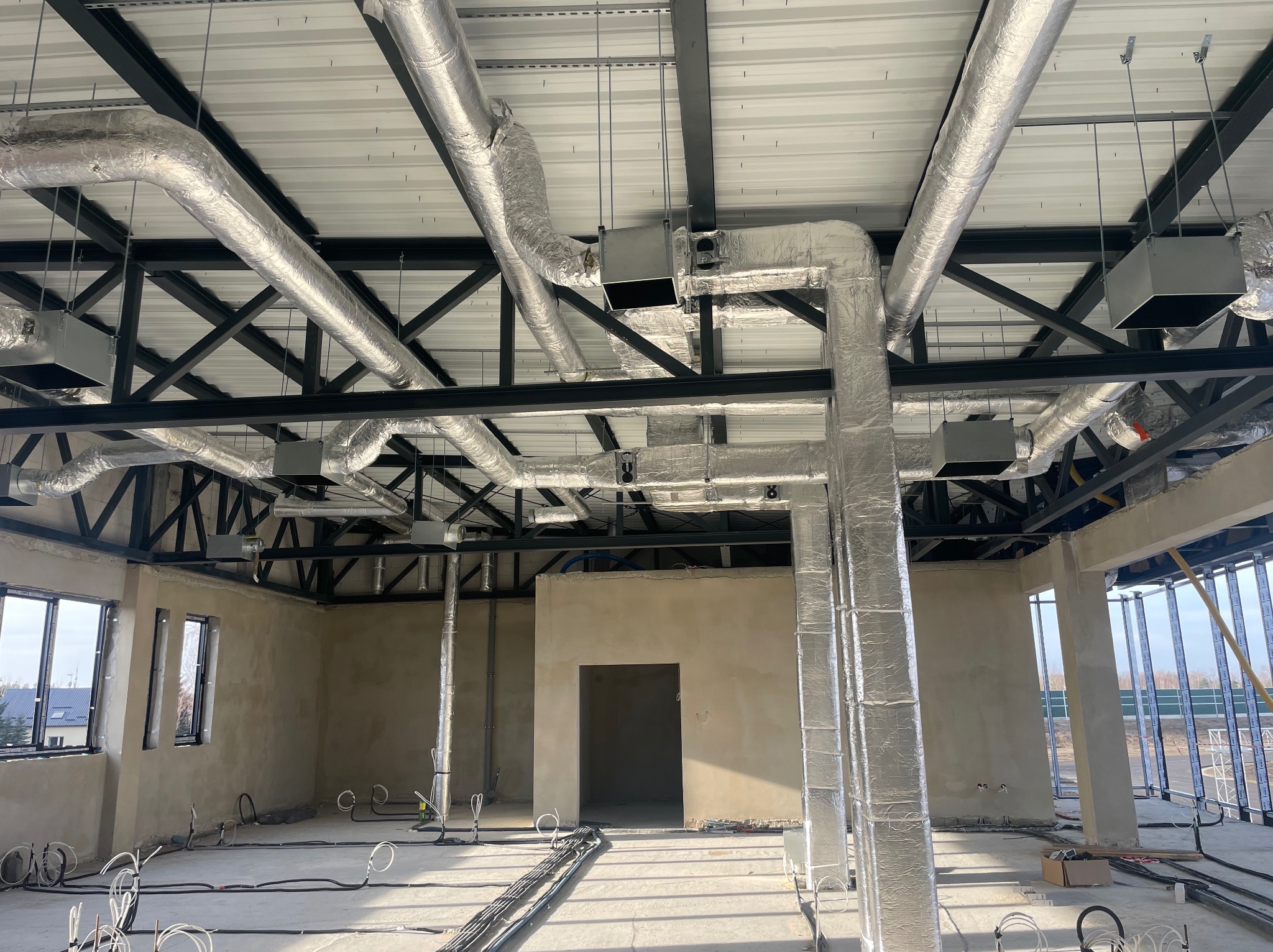 Wnętrze budynku w trakcie instalacji systemu wentylacji: widoczne kanały wentylacyjne owinięte folią aluminiową pod sufitem, czarne elementy konstrukcyjne dachu, surowy stan ścian i podłogi...