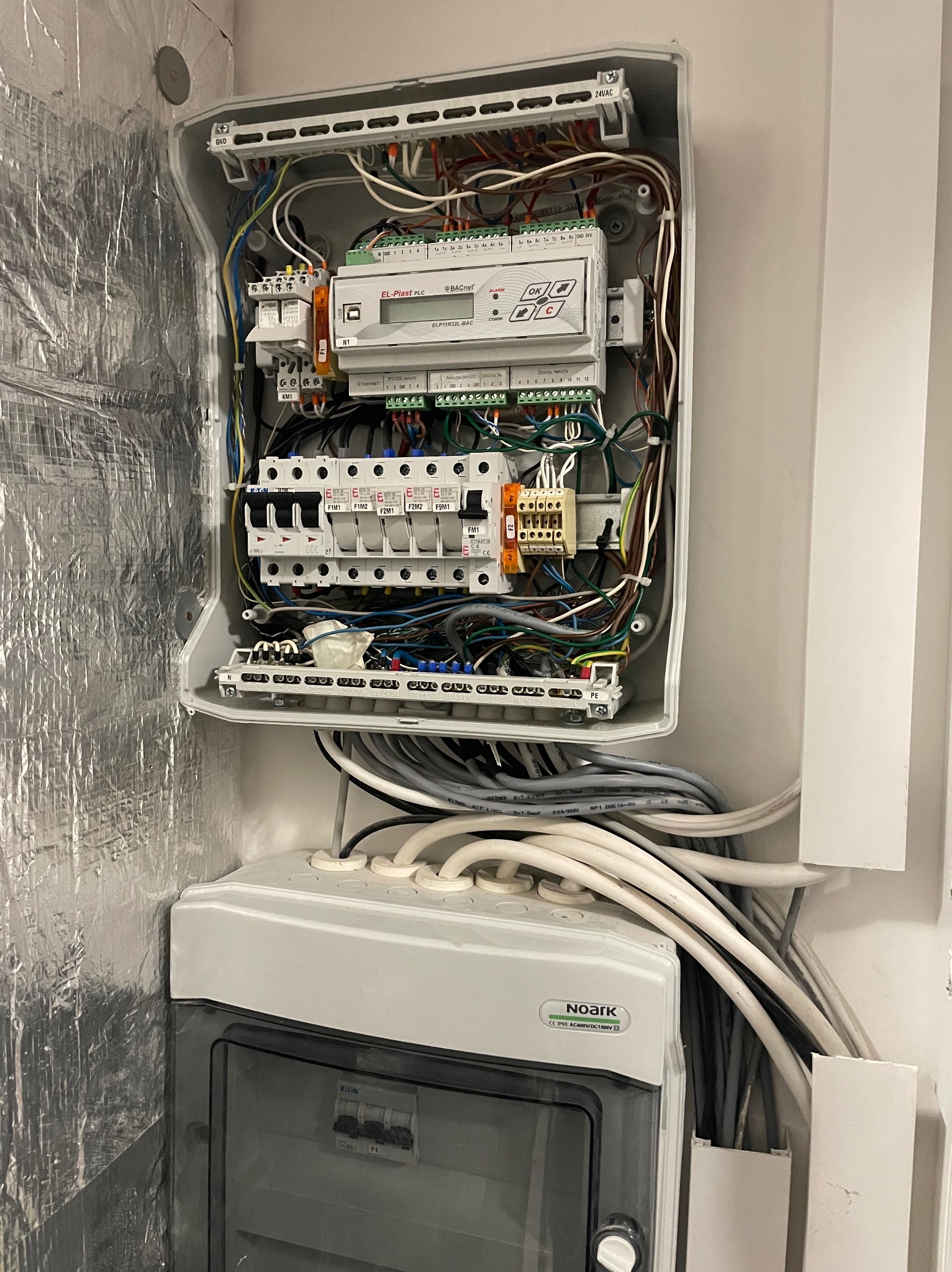 Otwarta skrzynka sterownicza z widocznymi przewodami, bezpiecznikami i modułem sterującym EL-Piast PLC na ścianie z izolacją aluminiową oraz dolna skrzynka Noark z widocznymi przewodami.