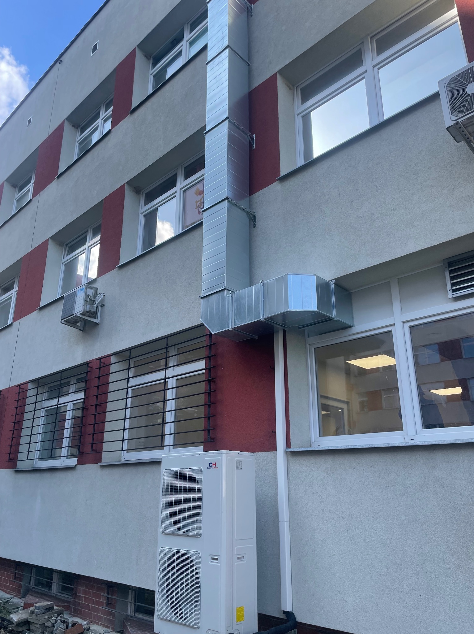 Zewnętrzna instalacja wentylacyjna z rurami i klimatyzator na elewacji budynku z czerwonymi akcentami, widoczne okna z kratami.