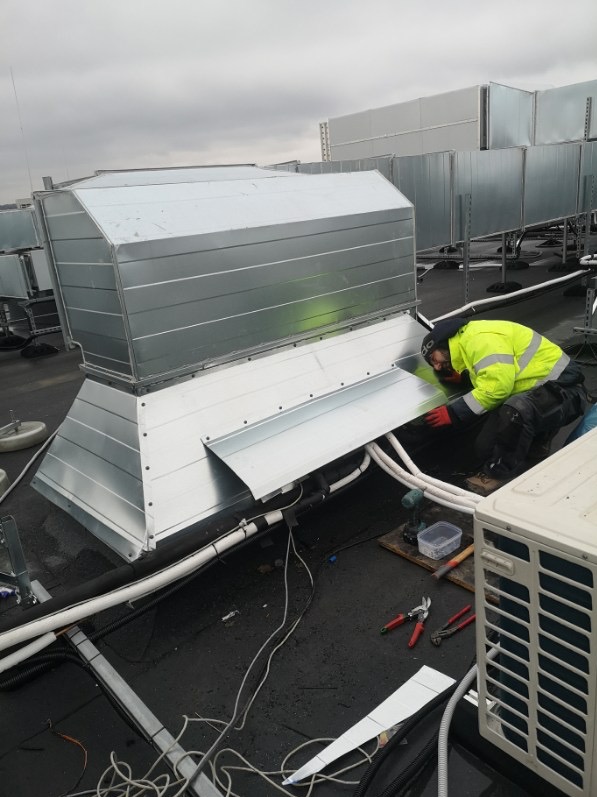 Pracownik w żółtej kurtce montuje metalowy kanał wentylacyjny na dachu budynku, widoczne narzędzia i kable.