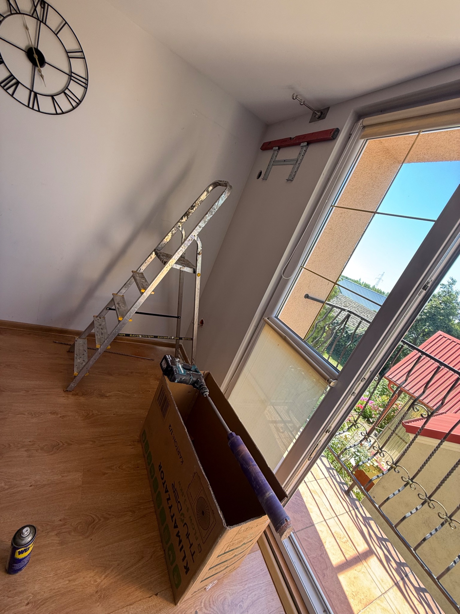 Wnętrze pokoju z drabiną, narzędziami i uchwytem na ścianie, prawdopodobnie podczas montażu klimatyzacji. Widoczny fragment okna i pudełko po klimatyzatorze.