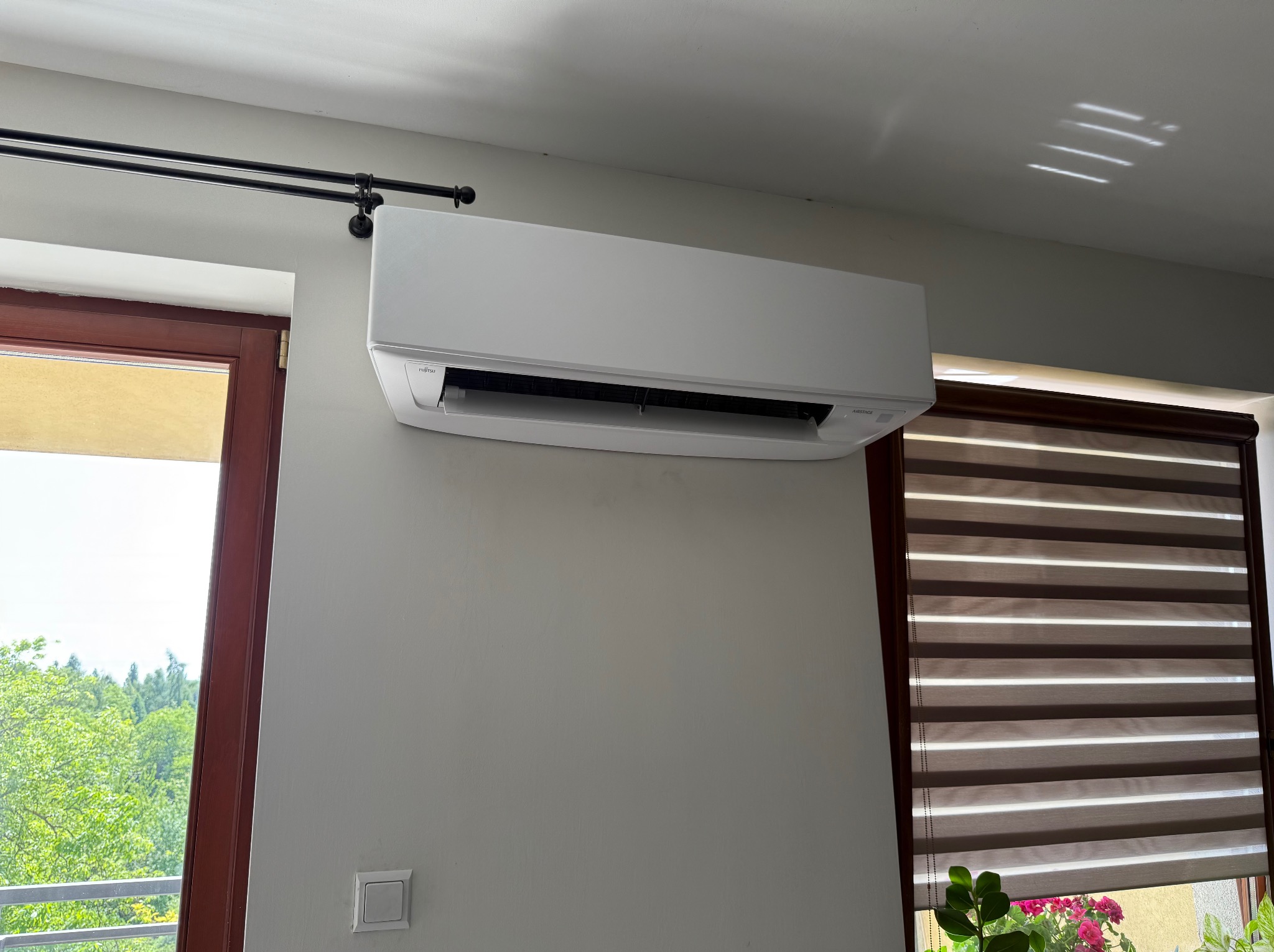 Biały klimatyzator Fujitsu Airstage zamontowany na ścianie w jasnym pokoju, widok z dołu, okno z roletami w tle.