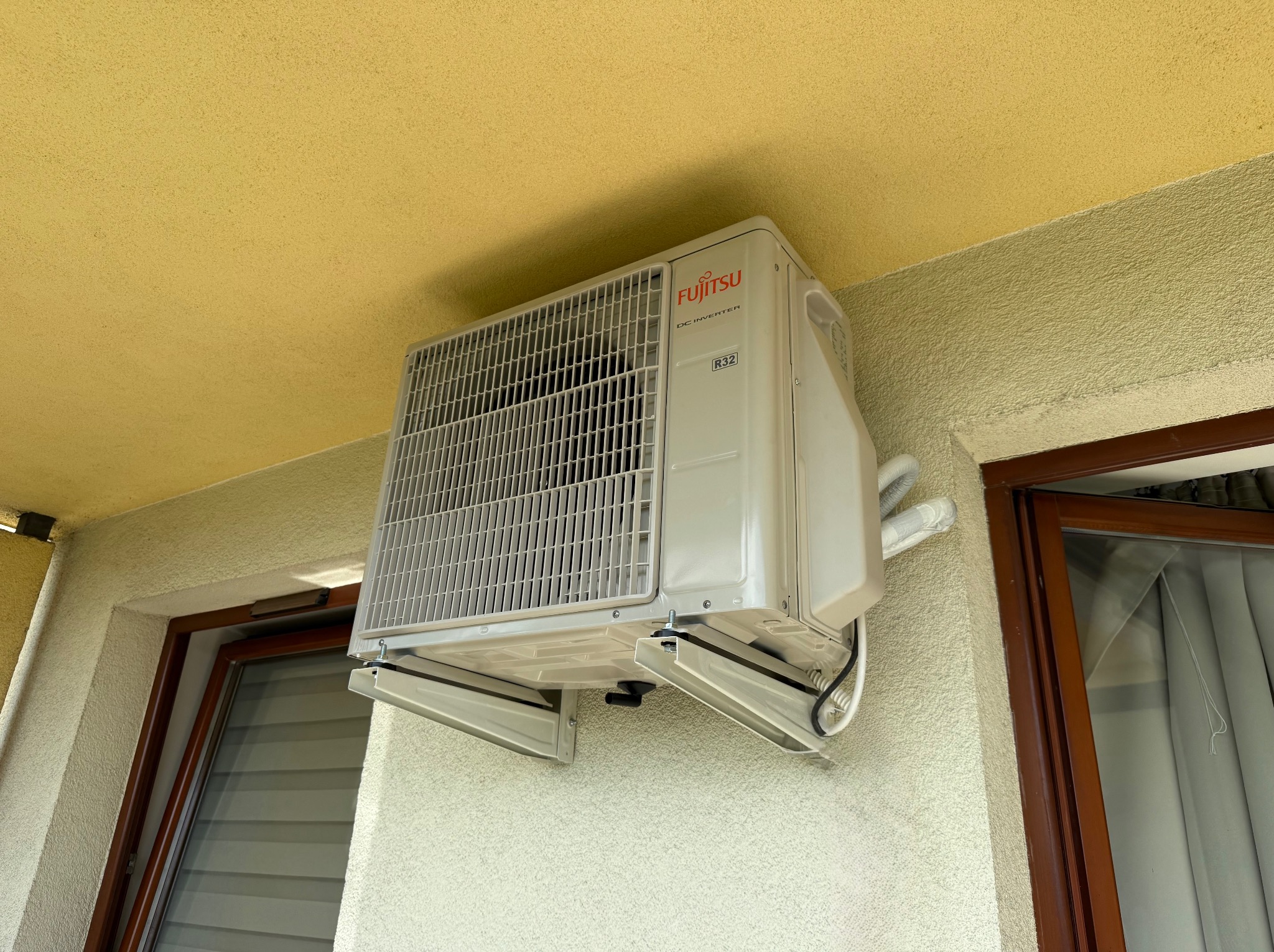 Zamontowany na ścianie, biały klimatyzator Fujitsu z widocznymi rurami i elementami montażowymi, zainstalowany nad oknem z brązową ramą. Żółty sufit w tle.
