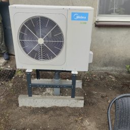 GREEN AIR POWER - Zewnętrzna jednostka pompy ciepła Midea zamontowana na betonowej podstawie z metalowym stelażem, widoczne krople deszczu, przy szarej ścianie budynku z oknem