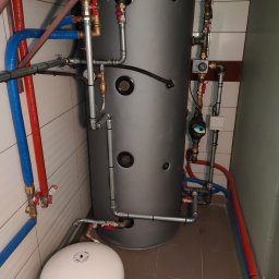 GREEN AIR POWER - Szary zbiornik buforowy ciepła z podłączonymi rurami miedzianymi, zaworami i pompą obiegową w kotłowni z białymi płytkami na ścianach i brązowymi na podłodze.