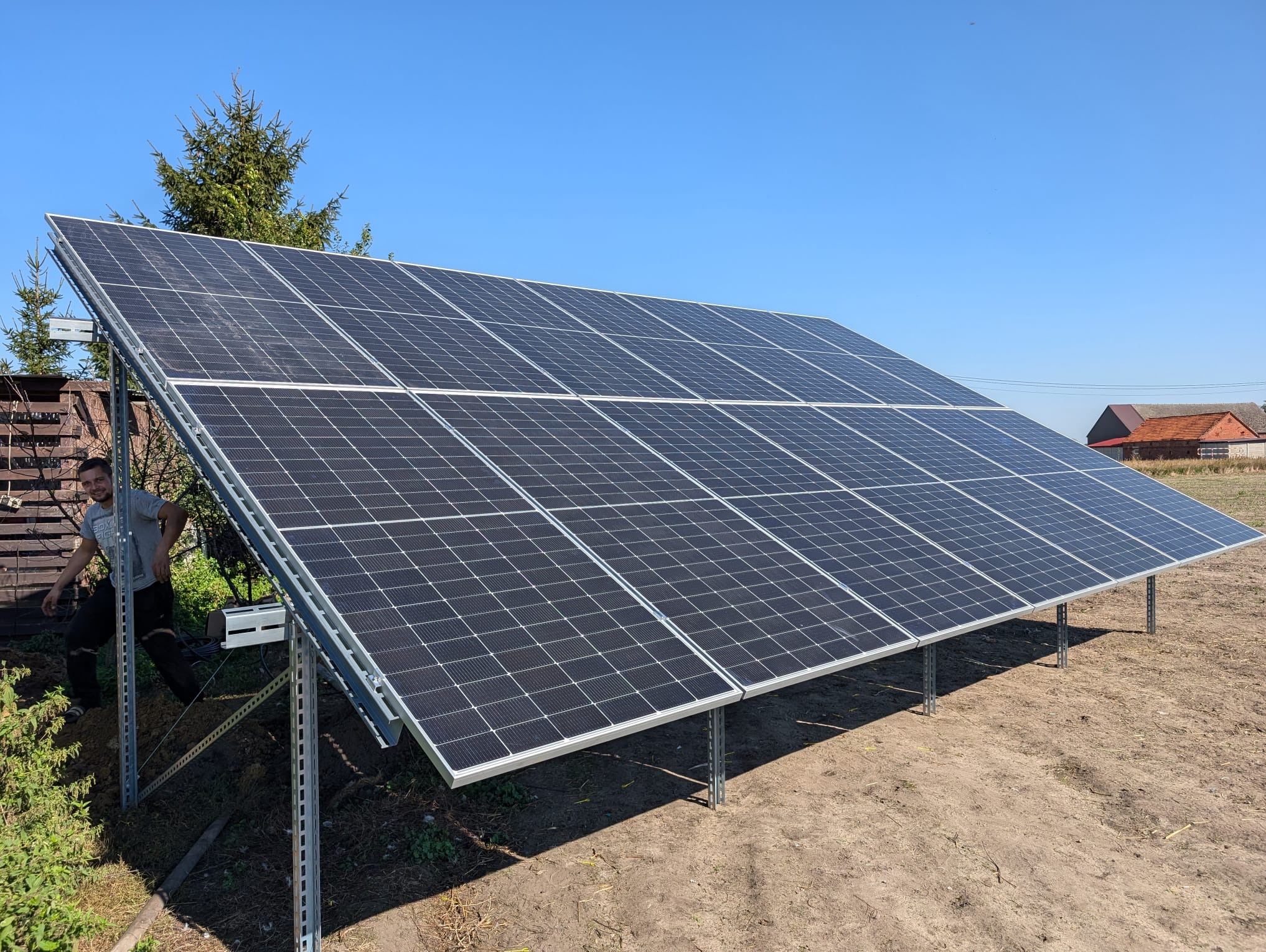 Zestaw paneli słonecznych na metalowej konstrukcji, widoczny instalator w szarej koszulce, tło: wiejski krajobraz i błękitne niebo.