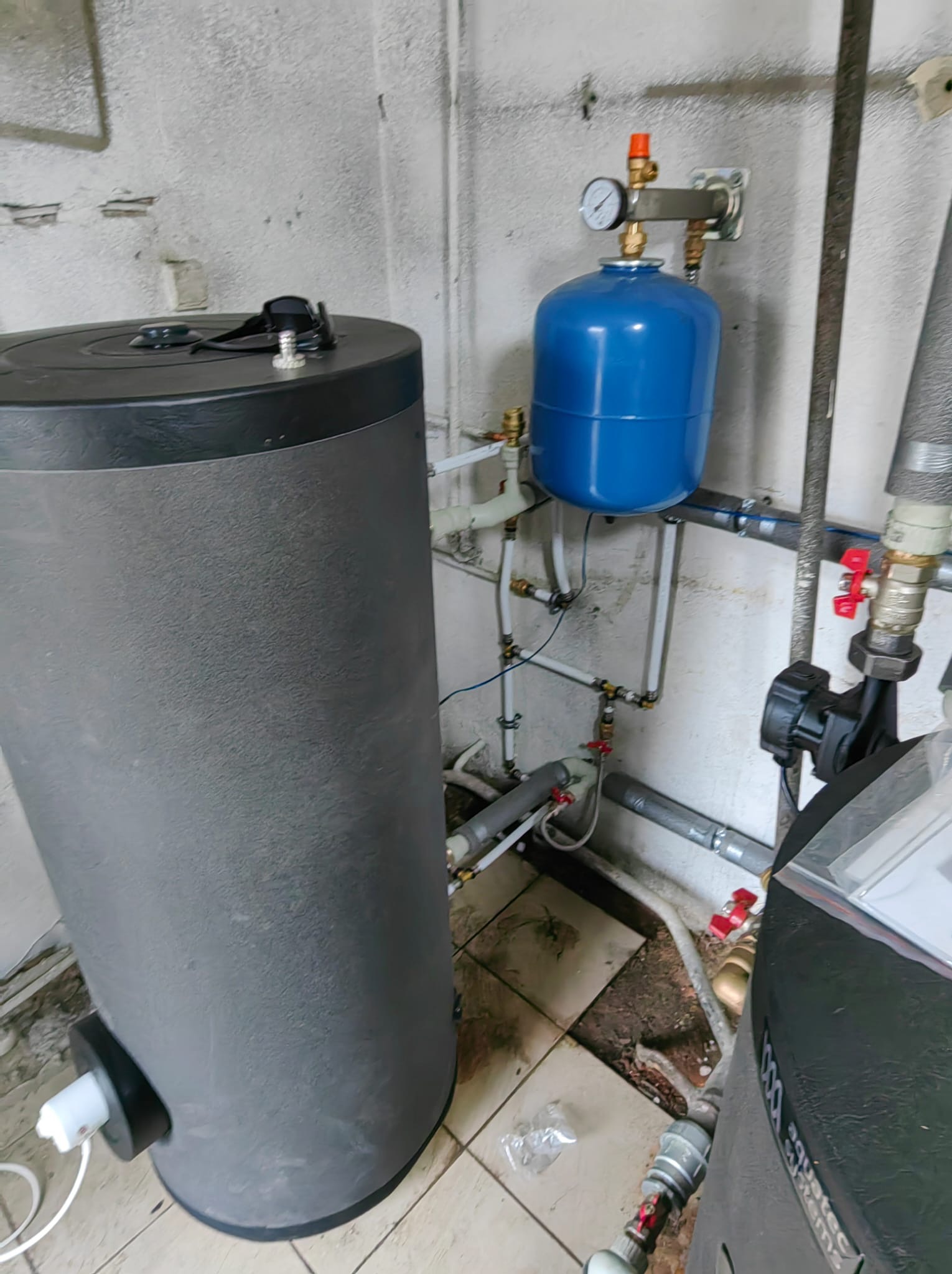 Szary zbiornik akumulacyjny ciepła z podłączoną niebieską membraną, manometrem i instalacją hydrauliczną w pomieszczeniu technicznym z widocznymi rurami i zaworami.
