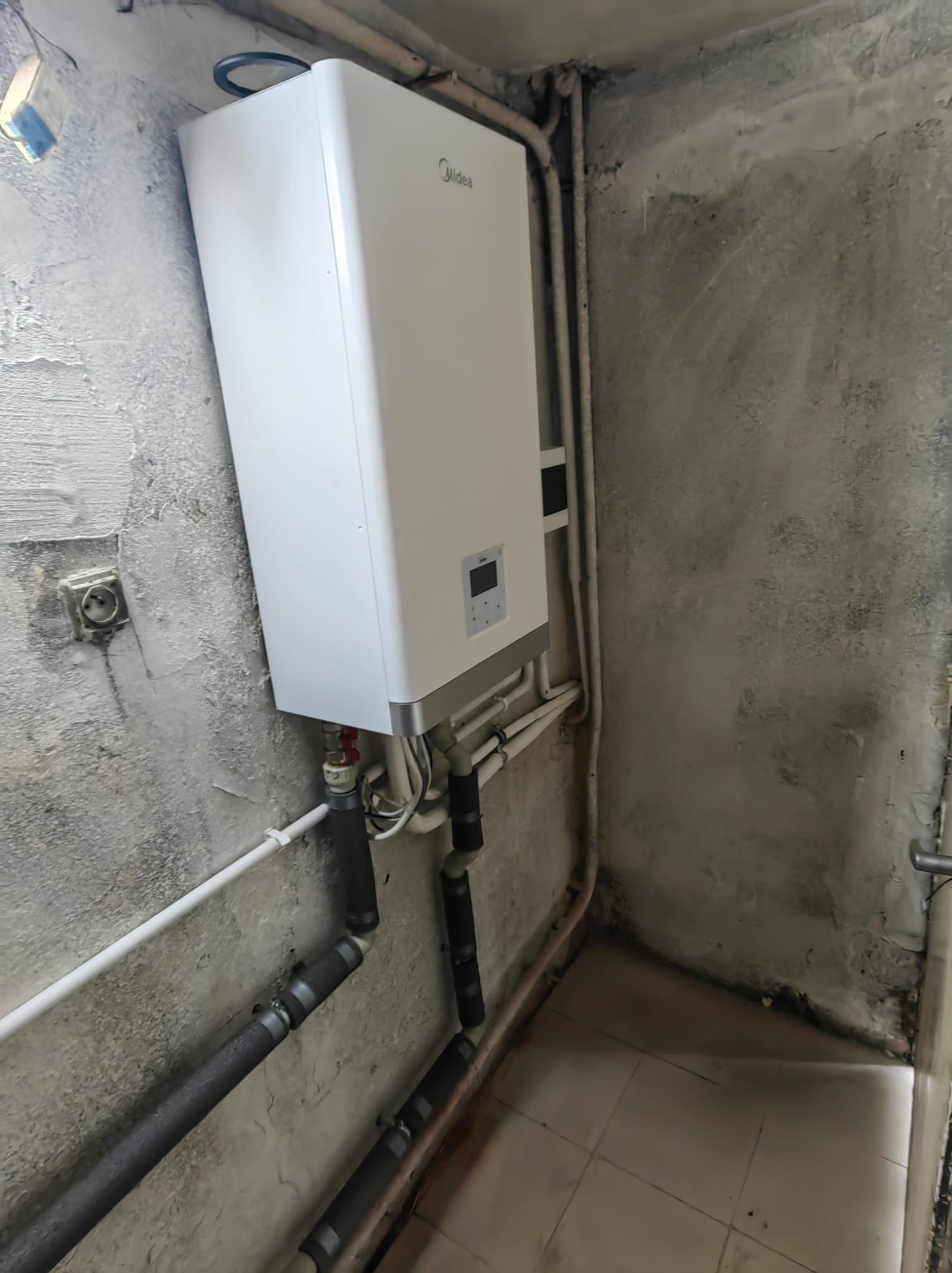 Biały panel grzewczy Midea zamontowany na szarej, surowej ścianie z widocznymi rurami i gniazdkiem elektrycznym.