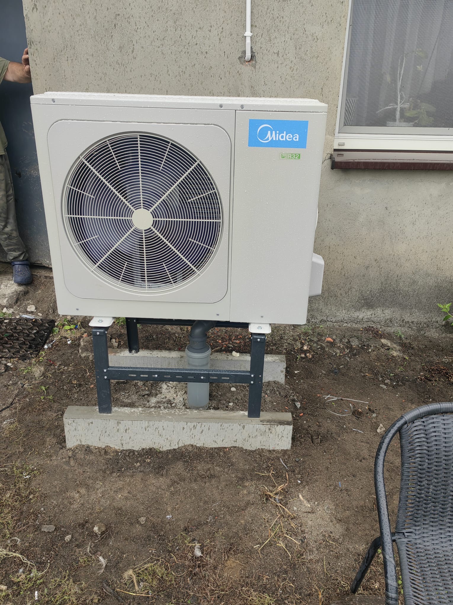 Zewnętrzna jednostka pompy ciepła Midea zamontowana na betonowej podstawie z metalowym stelażem, widoczne krople deszczu, przy szarej ścianie budynku z oknem