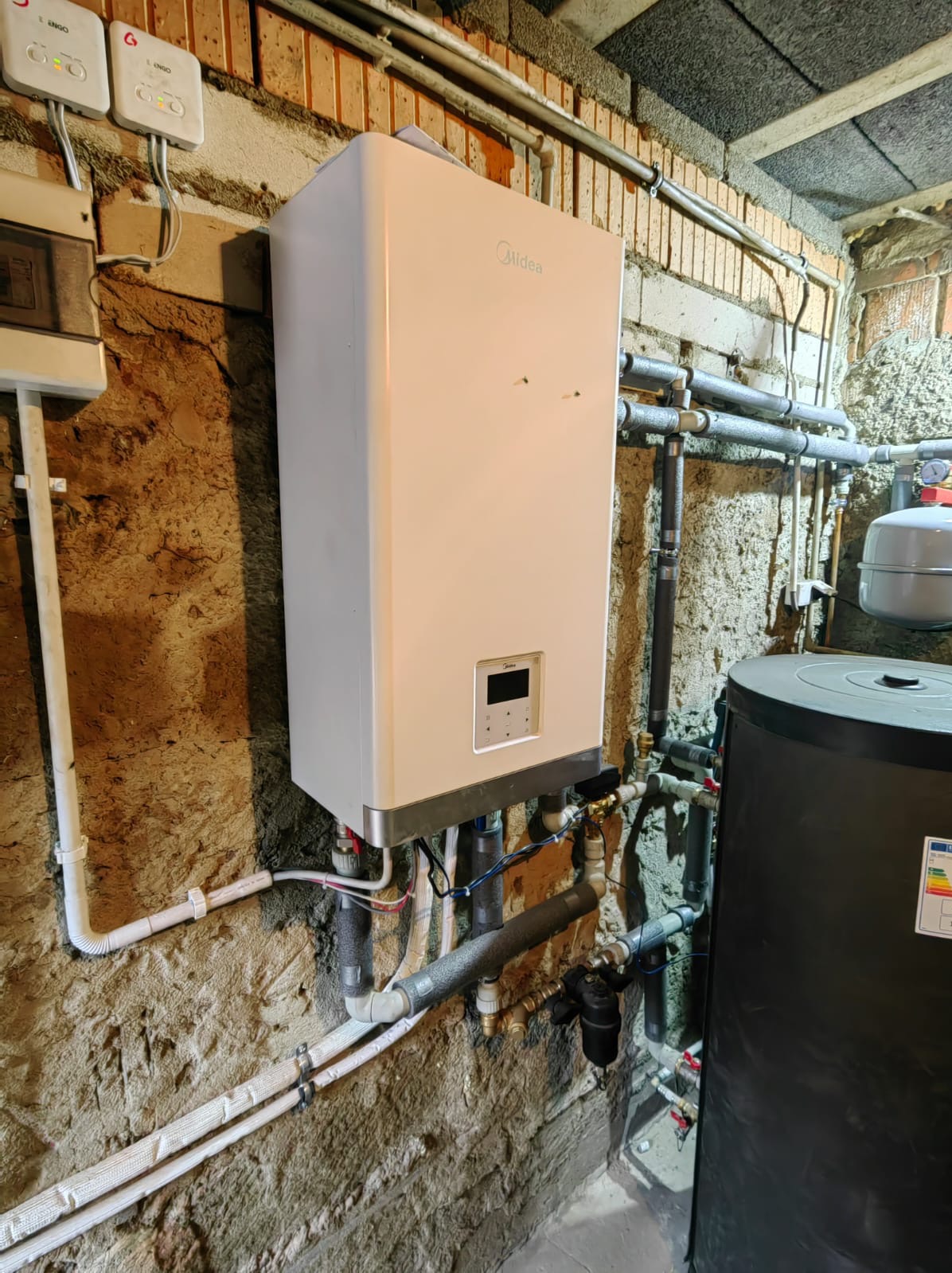 Biały, wiszący moduł fotowoltaiczny Midea z podłączeniami rur na tle surowej, ceglanej ściany piwnicy, widoczne liczniki i zbiornik buforowy.