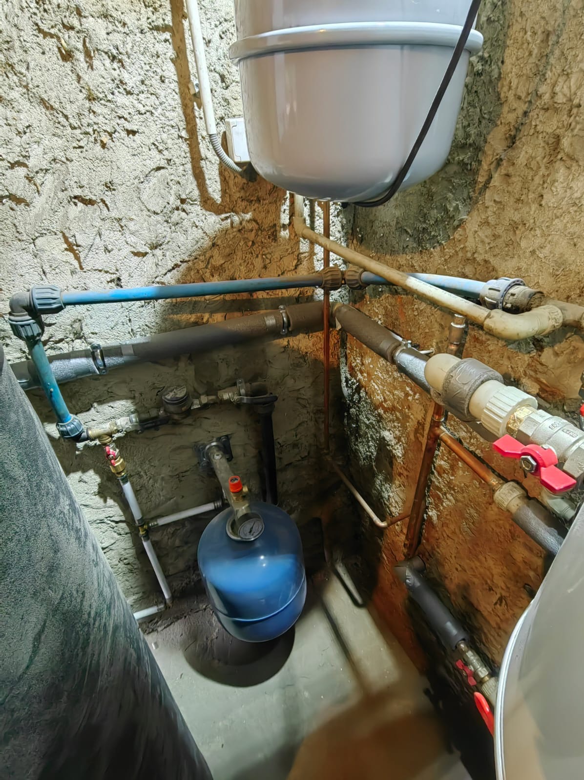 Instalacja hydrauliczna w piwnicy w Lusówku, widoczne rury miedziane, plastikowe, zawory, manometr, zbiornik ciśnieniowy i bojler na tle surowych ścian.