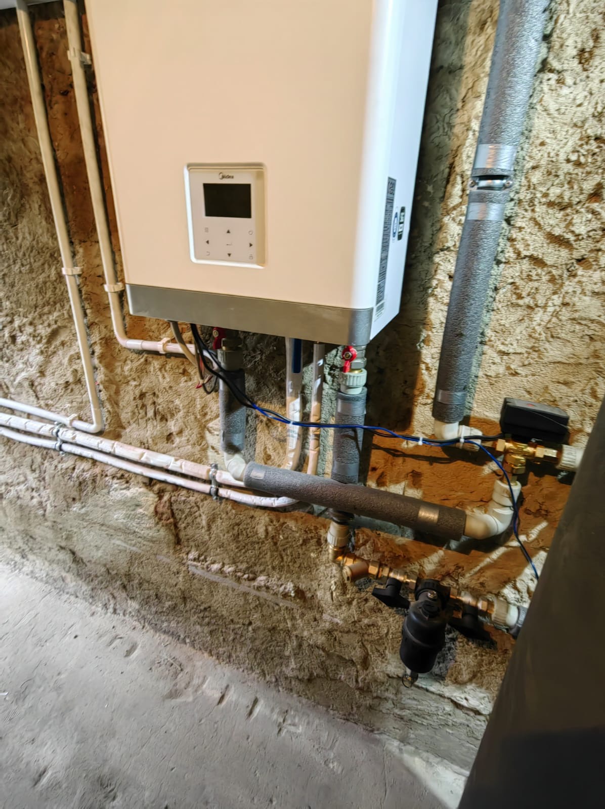 Biały piec grzewczy Midea zamontowany na szorstkiej, nierównej ścianie z widocznymi rurami i przewodami.