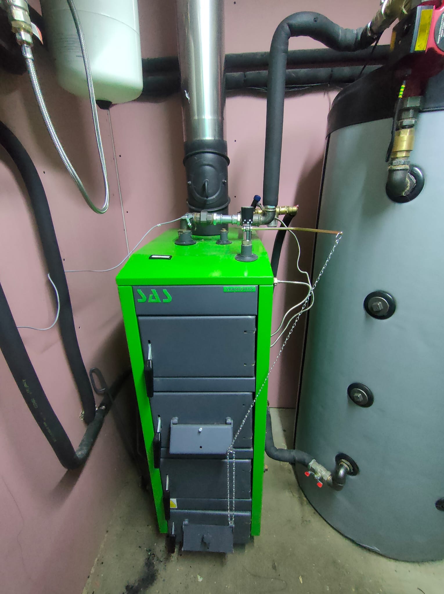 Kotłownia z kotłem na pellet marki SAS w kolorze zielonym i szarym, obok zbiornik akumulacyjny, widoczne rury i instalacja grzewcza.