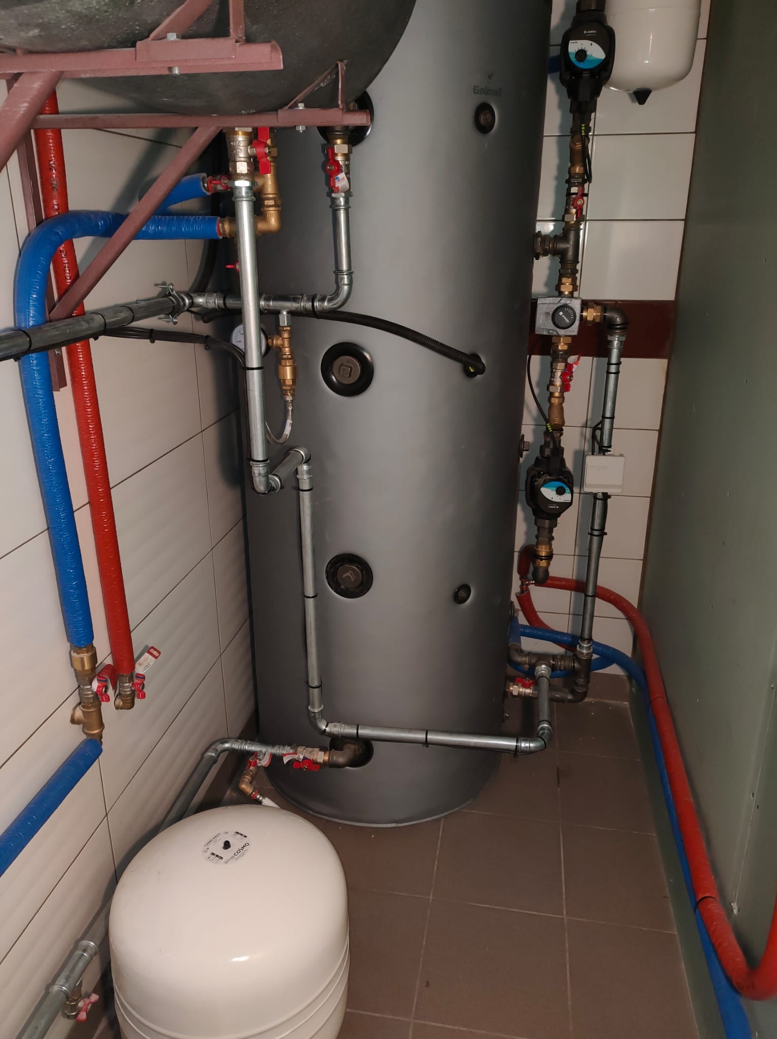 Szary zbiornik buforowy ciepła z podłączonymi rurami miedzianymi, zaworami i pompą obiegową w kotłowni z białymi płytkami na ścianach i brązowymi na podłodze.