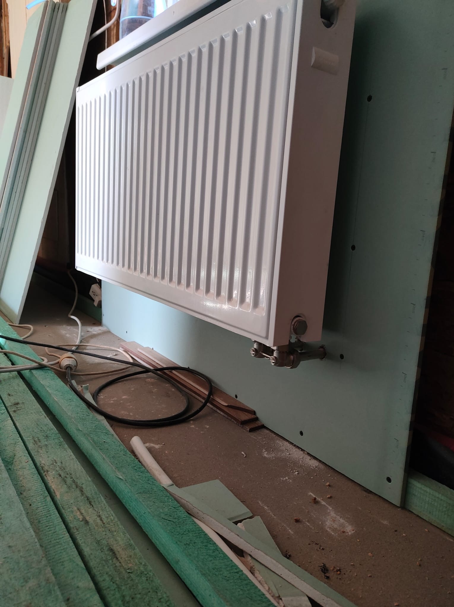 Biały grzejnik panelowy zamontowany na ścianie pokrytej płytą gipsowo-kartonową, widoczne przewody i elementy montażowe, w tle i na podłodze resztki materiałów budowlanych.