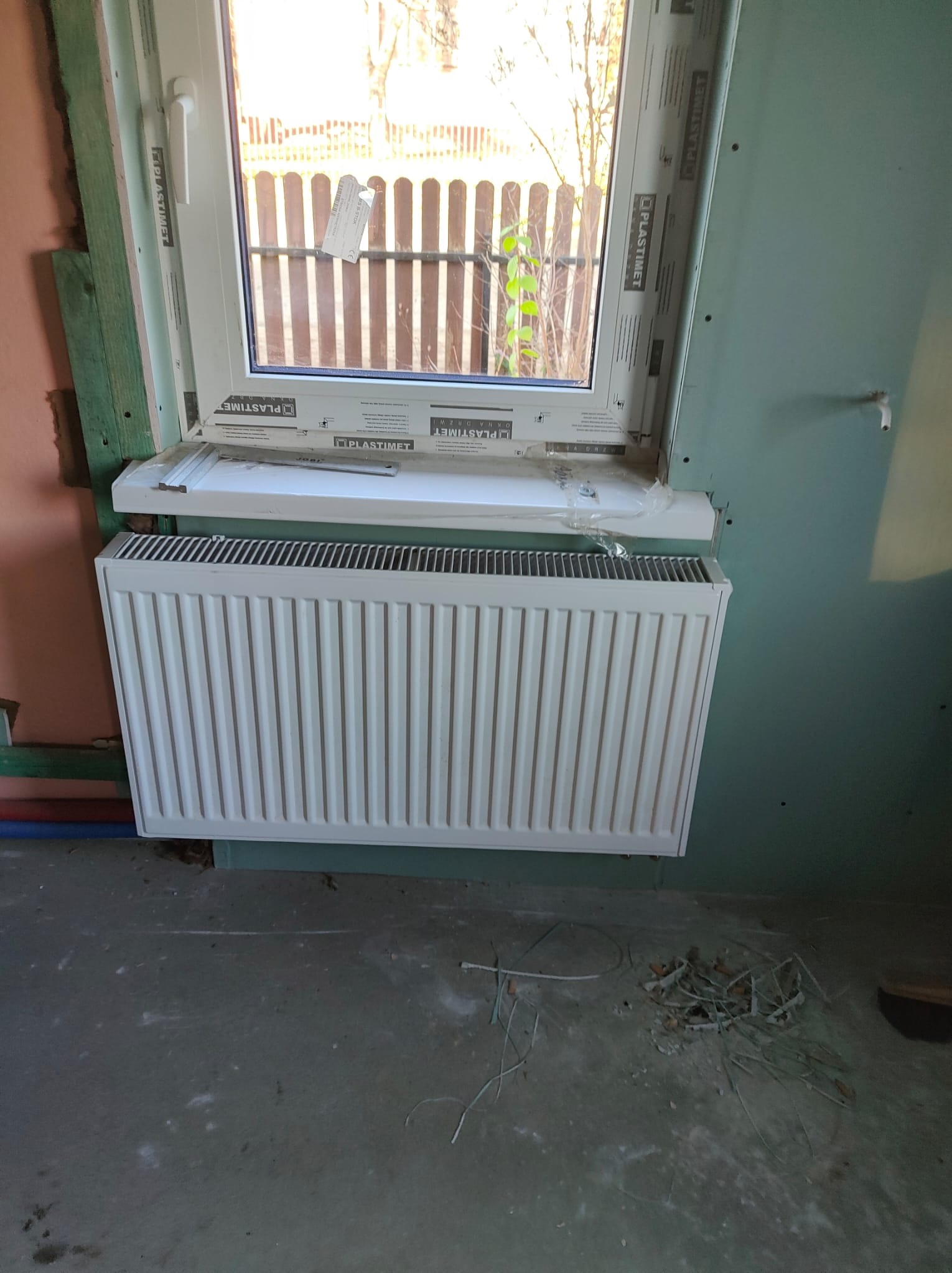 Biały grzejnik panelowy zamontowany pod oknem z PCV w trakcie remontu, widoczne surowe ściany i bałagan na podłodze.