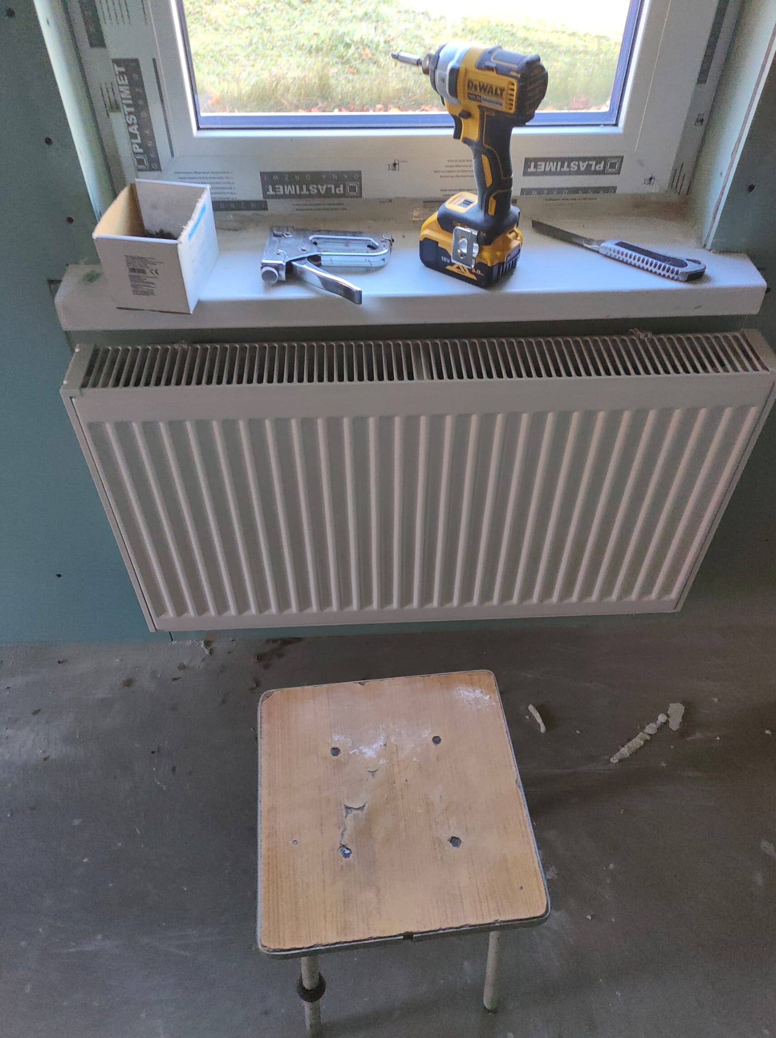 Montaż grzejnika pod oknem z użyciem wkrętarki DeWalt, zszywacza tapicerskiego i nożyka do tapet, widoczny stołek roboczy.