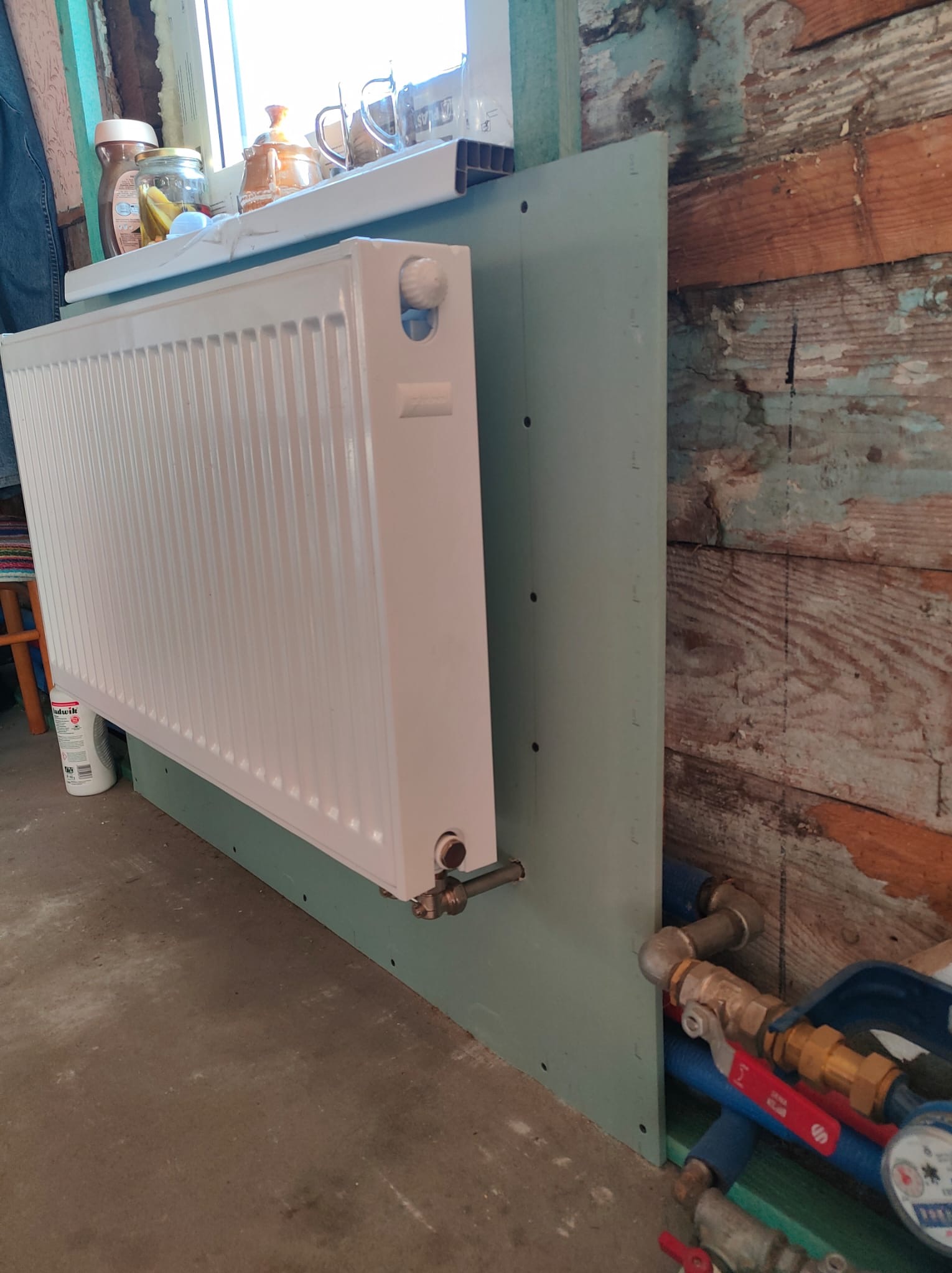 Zamontowany biały grzejnik panelowy na tle ściany z płyty gipsowej i fragmentu drewnianej ściany z odpryskującą farbą, widoczne rury doprowadzające wodę i elementy instalacji grzewczej.